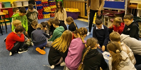 Powiększ grafikę: Dwie grupy dzieci siedzą na dywanie. Nauczycielka pochyla się w ich stronę pokazując na plakaty, które leżą na podłodze. Za nimi stoi granatowa tablica informacyjna dotycząca strojów kociewskich.