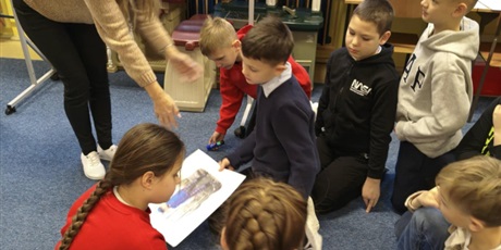 Powiększ grafikę: Grupa dzieci siedzi na dywanie. Nauczycielka pochyla się w ich stronę pokazując na plakaty, które trzymają dzieci. Za nimi stoi granatowa tablica informacyjna dotycząca strojów kociewskich.