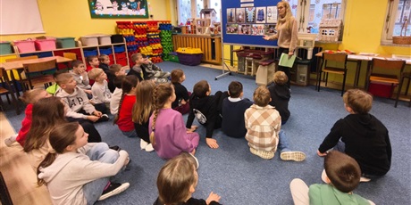 Powiększ grafikę: Grupa dzieci siedzą na dywanie i wpatruje się w nauczycielkę, która do nich mówi. W centralnym miejscu stoi granatowa tablica informacyjna dotycząca strojów kociewskich.