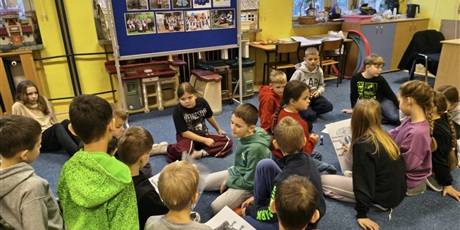 Powiększ grafikę: Dwie grupy dzieci siedzą na dywanie. W centralnym miejscu stoi granatowa tablica informacyjna dotycząca strojów kociewskich.