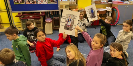 Powiększ grafikę:  Grupa dzieci siedzi na dywanie w świetlicy. Dwie dziewczynki i dwóch chłopców trzymają uniesione do góry plakaty z postacią kobiety i mężczyzny ubranych w stroje kociewskie.