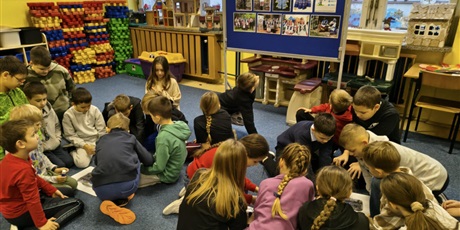 Powiększ grafikę: Dwie grupy dzieci siedzą w dwóch kręgach na dywanie. Dzieci pochylają się nad plakatami, które leżą między nimi. Za nimi stoi granatowa tablica informacyjna dotycząca strojów kociewskich.