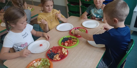 Powiększ grafikę: Dzieci siedzą przy stoliku. Przed nimi są talerzyki, na których leżą pokrojone w kostkę owoce. Dziewczynki nabijają owoce na pałeczki do szaszłyków.