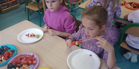 Powiększ grafikę: Dzieci siedzą przy stoliku. Przed nimi są talerzyki, na których leżą pokrojone w kostkę owoce. Dziewczynki nabijają owoce na pałeczki do szaszłyków.