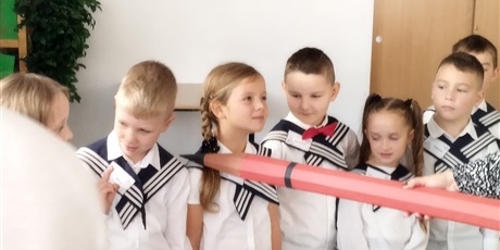 Powiększ grafikę: Dziewczynka jest pasowana na zerówkowicza. Na ramię położono jej duży ołówek i pasowano na zerówkowicza. Obok stoją pozostali uczniowie i czekają na swoją kolej.