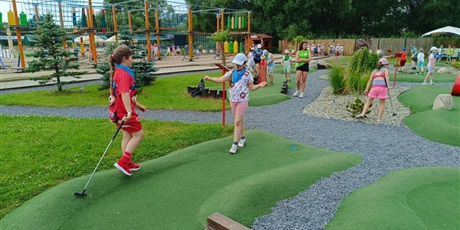 Powiększ grafikę: Na placu zabaw, na świeżym powietrzu dzieci grają w mini golfa trzymając w dłoniach kije golfowe. Na pierwszym planie  zielone ścieżki do gry.