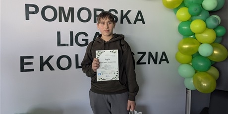 Powiększ grafikę: Laureat konkursu z dyplomem w ręku na tle ścianki z napisem „Pomorska Liga Ekologiczna” ozdobionej zielonymi balonami.