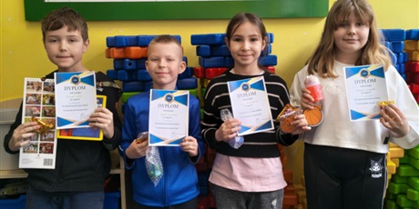 Powiększ grafikę: Grupa dzieci pozuje do zdjęcia. W rękach trzymają dyplomy i nagrody za udział w konkursie. Za nimi- na ścianie  wisi zielona tablica z zimową dekoracją.