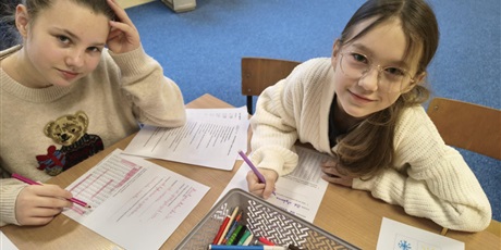 Powiększ grafikę: Dwie dziewczynki siedzą przy stole i patrzą w obiektyw. Na stole leżą kartki z zadaniami do rozwiązania.