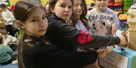 Powiększ grafikę: Cztery dziewczynki pozują do zdjęcia. Wrzucają kartki z rozwiązanymi zadaniami do niebieskiego pudełka z napisem QUIZ WIEDZY O KALENDARZU.