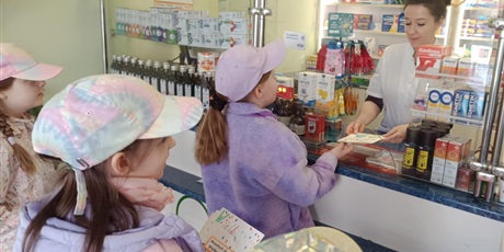 Powiększ grafikę: Dziewczynka wręcza kartkę wielkanocną pani farmaceutce w aptece. Za nią stoją dwie inne dziewczynki. Wszystkie mają na głowie czapki