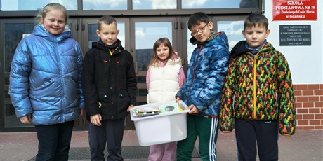 Powiększ grafikę: Dwie dziewczynki i trzech chłopców stoją przed wejściem do szkoły. Dwóch chłopców w środku trzyma białe pudełko z kartkami wielkanocnymi.