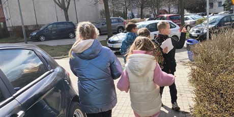 Powiększ grafikę: Grupa dzieci widoczna tyłem idzie chodnikiem. Obok znajdują się samochody.