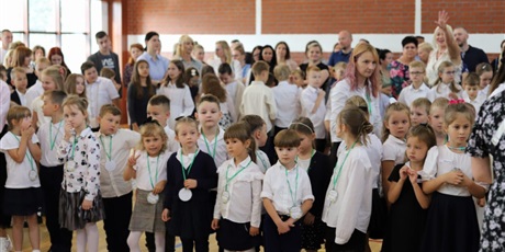 Powiększ grafikę: Grupa uczniów w strojach galowych znajduje się w sali gimnastycznej. Dzieci spoglądają na fotografa  oraz inne dzieci.