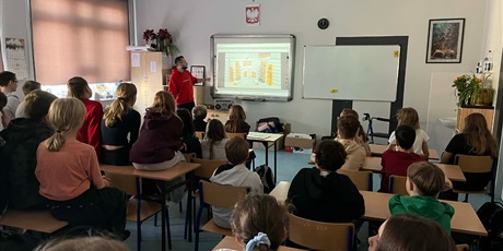 Powiększ grafikę: Na zdjęciu widać uczniów słuchających opowieści pracownika firmy Lotte Wedel. Uczniowie siedzą na krzesełkach i na ławkach. Jakub pokazuje prezentację na tablicy.