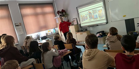 Powiększ grafikę: Na zdjęciu widać uczniów słuchających opowieści pracownika firmy Lotte Wedel. Uczniowie siedzą na krzesełkach i na ławkach. Jakub pokazuje prezentację na tablicy.