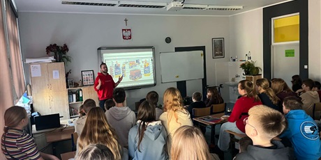 Powiększ grafikę: Na zdjęciu widać uczniów słuchających opowieści pracownika firmy Lotte Wedel. Uczniowie siedzą na krzesełkach i na ławkach. Jakub pokazuje prezentację na tablicy.