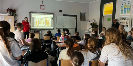 Powiększ grafikę: Na zdjęciu widać uczniów słuchających opowieści pracownika firmy Lotte Wedel. Uczniowie siedzą na krzesełkach i na ławkach. Jakub pokazuje prezentację na tablicy.