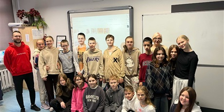 Powiększ grafikę: Uczniowie z klasy szóstej pozują do zdjęcia z Jakubem Szczepankowskim (mężczyzna z brodą, w czerwonej bluzie). W tle widać klasę oraz tablicę z prezentacją, o której mówił wcześniej Jakub.