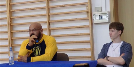 Powiększ grafikę: Michał Kamrowski w żółtej bluzie mówiący przez mikrofon. Obok siedzi uczeń.