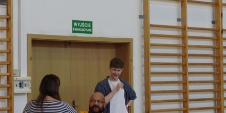 Powiększ grafikę: Michał Kamrowski w żółtej bluzie odbierający mikrofon.  Kobieta podaje mikrofon. W tle stoi uczeń. 