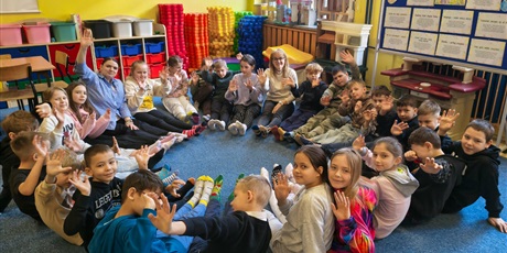 Powiększ grafikę: Grupa dzieci wraz z nauczycielkami siedzi w kręgu na dywanie. Wszyscy zwróceni są w stronę obiektywu. Na stopach mają kolorowe skarpetki.