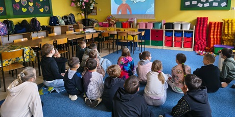 Powiększ grafikę: Grupa dzieci siedzi na dywanie tyłem do obiektywu. Wszyscy patrzą na ścienny ekran, na którym wyświetlony jest film.