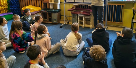 Powiększ grafikę: Grupa dzieci siedzi na dywanie w świetlicy. Wszyscy zwróceni są w stronę nauczycielki, która stoi obok granatowej tablicy i czyta dzieciom opowiadanie.