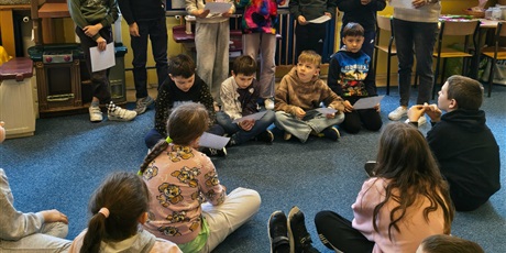 Powiększ grafikę: Dziesięcioro dzieci stoi lub siedzi na tle granatowej tablicy informacyjnej. Wszyscy trzymają w dłoni białe kartki, z których czytają pozostałym uczniom informacje. Obok stoi nauczycielka.