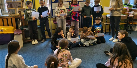 Powiększ grafikę: Dziesięcioro dzieci stoi lub siedzi na tle granatowej tablicy informacyjnej. Wszyscy trzymają w dłoni białe kartki, z których czytają pozostałym uczniom informacje. Obok stoi nauczycielka.