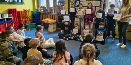 Powiększ grafikę: Dziesięcioro dzieci stoi lub siedzi na tle granatowej tablicy informacyjnej. Wszyscy trzymają w dłoni białe kartki, z których czytają pozostałym uczniom informacje. Obok stoi nauczycielka.