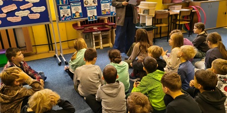 Powiększ grafikę: Grupa dzieci siedzi na dywanie w świetlicy zwrócona w kierunku nauczycielki, która czyta opowiadanie o życzliwości. Z tyłu znajdują się  dwie granatowe tablice z informacjami dotyczącymi Światowego Dn