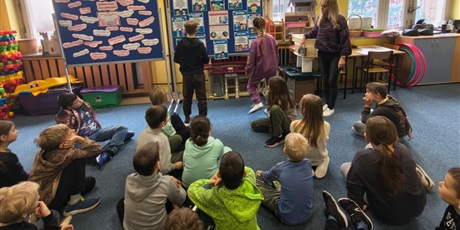 Powiększ grafikę: Grupa dzieci siedzi na dywanie w świetlicy zwrócona tyłem do obiektywu. Dzieci patrzą na dziewczynkę i chłopca, którzy stoją i czytają informacje z tablicy na temat Światowego Dnia Życzliwości i Pozdr