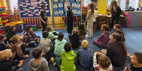 Powiększ grafikę: Grupa dzieci siedzi na dywanie w świetlicy zwrócona tyłem do obiektywu. Dzieci patrzą na dziewczynkę i dwóch chłopców,  którzy stoją i czytają informacje z tablicy na temat Światowego Dnia Życzliwości