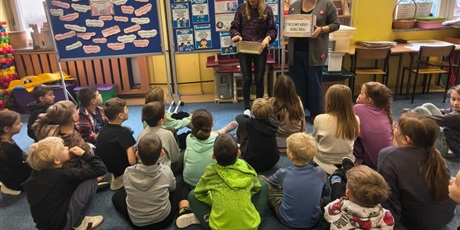 Powiększ grafikę: Grupa dzieci siedzi na dywanie w świetlicy zwrócona tyłem do obiektywu. Dzieci patrzą na dwie nauczycielki, które trzymają w rękach pudełko "Życzliwe Hasło- Podaj Dalej". Z tyłu znajdują się dwie tabl