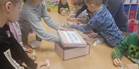 Powiększ grafikę: Grupa dzieci stoi lub siedzi przy stole w świetlicy. Dzieci piszą na kolorowych karteczkach życzliwe hasła i pozdrowienia.