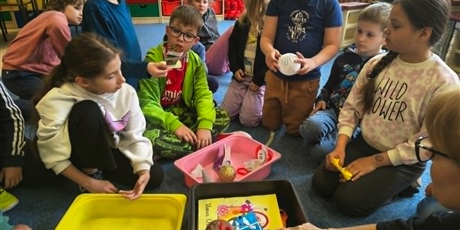 Powiększ grafikę: Grupa dzieci wraz z nauczycielkami siedzą na dywanie. Z kolorowych pojemników przekładają, przygotowane przez siebie, przedmioty do Kapsuły Czasu.