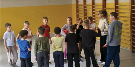 Powiększ grafikę: Dwie grupy dzieci stoją na boisku na przeciw siebie. Na środku stoi nauczycielka.