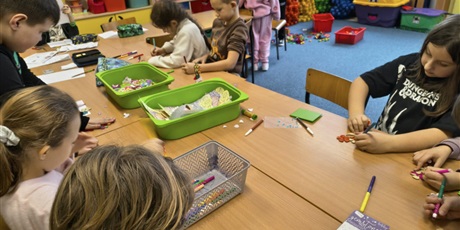 Powiększ grafikę: Grupa dzieci siedzi przy stole i wykonuje upominki dla babci i dziadka. Na stole leżą zielone i szare pojemniki z akcesoriami do dekoracji.