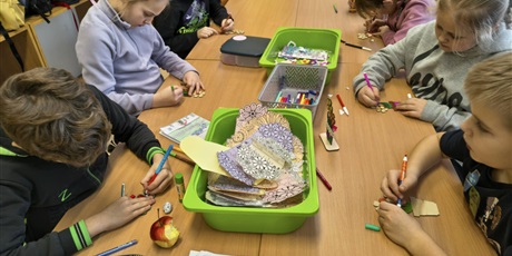 Powiększ grafikę: Grupa dzieci siedzi przy stole i wykonuje upominki dla babci i dziadka. Na stole leżą zielone i szare pojemniki z akcesoriami do dekoracji.