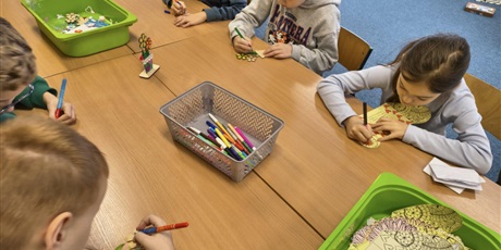 Powiększ grafikę: Grupa dzieci siedzi przy stole i wykonuje upominki dla babci i dziadka. Na stole leżą zielone i szare pojemniki z akcesoriami do dekoracji.