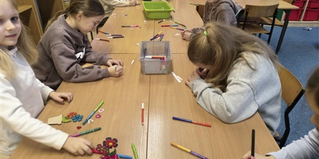 Powiększ grafikę: Grupa dzieci siedzi przy stole i wykonuje upominki dla babci i dziadka. Na stole leżą zielone i szare pojemniki z akcesoriami do dekoracji.