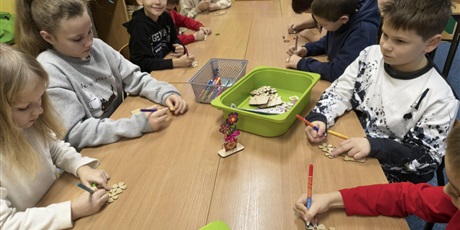 Powiększ grafikę: Grupa dzieci siedzi przy stole i wykonuje upominki dla babci i dziadka. Na stole leżą zielone i szare pojemniki z akcesoriami do dekoracji.