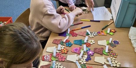 Powiększ grafikę: Dwie dziewczynki siedzą przy stole i wykonuje upominki dla babci i dziadka. Jedna dziewczynka stoi.  Na stole leżą kolorowe kwiatki.