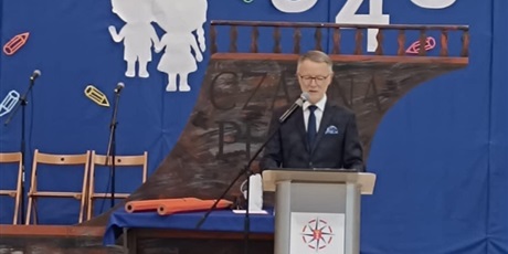 Powiększ grafikę: Mężczyzna w granatowym garniturze mówi do mikrofonu. Stoi przy mównicy na tle rufy statku „Czarna perła”.