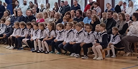 Powiększ grafikę: Na ławeczce siedzą dzieci w biało-czarnych odświętnych strojach i kołnierzach marynarskich. Za nimi siedzą dorośli.