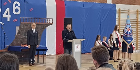 Powiększ grafikę: Na tle rufy statku „Czarna perła” stoi mężczyzna w granatowym garniturze. Drugi mężczyzna w ciemnej marynarce mówi do mikrofonu.