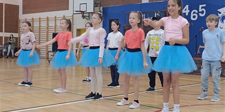 Powiększ grafikę: Uśmiechnięte dziewczynki w niebieskich spódniczkach i kolorowych bluzeczkach wykonują układ choreograficzny.