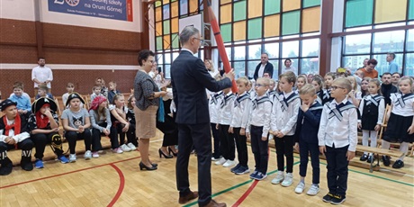 Powiększ grafikę: Mężczyzna w granatowym garniturze trzyma w dłoni dużą czerwoną kredkę. Przed nim stoją chłopcy w białych koszulach i marynarskich kołnierzach.