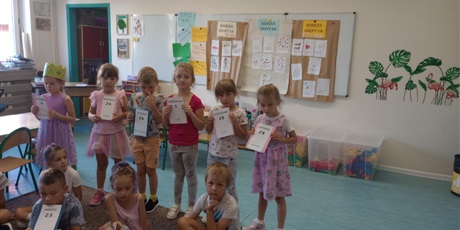Powiększ grafikę: Grupa dzieci sześcioletnich stoi na dywanie, każde dziecko trzyma w rękach swoją datę urodzin. Dni ułożone są chronologicznie.